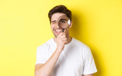 Close-up of young man looking through magnifying glass and smiling, searching something, standing over yellow background.